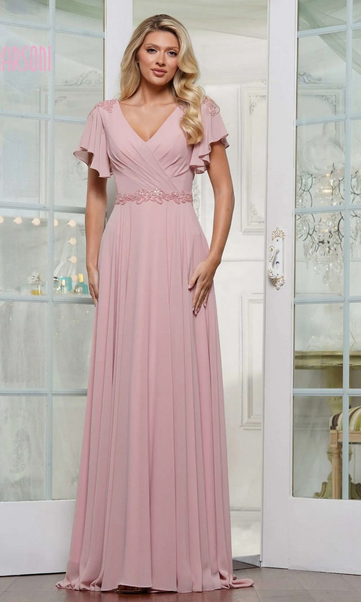 A woman stands indoors in the Marsoni M328 long formal dress in rose, featuring chiffon fabric, flutter sleeves, a V-neckline, lace detail at the shoulders, and a beaded waistband for an elegant look.